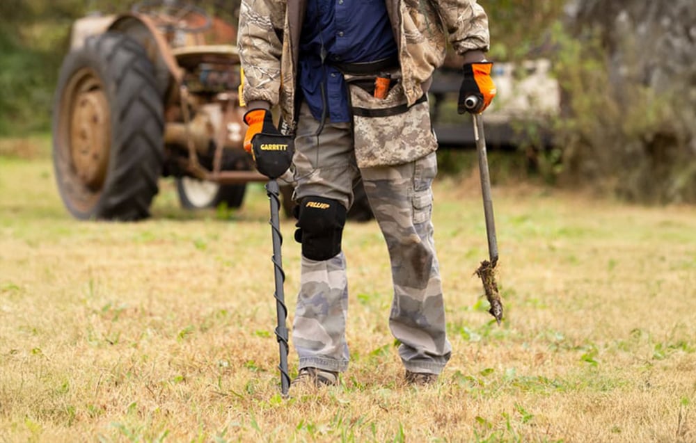 A person in camouflage clothing uses a metal detector in a field, with a tractor in the background.