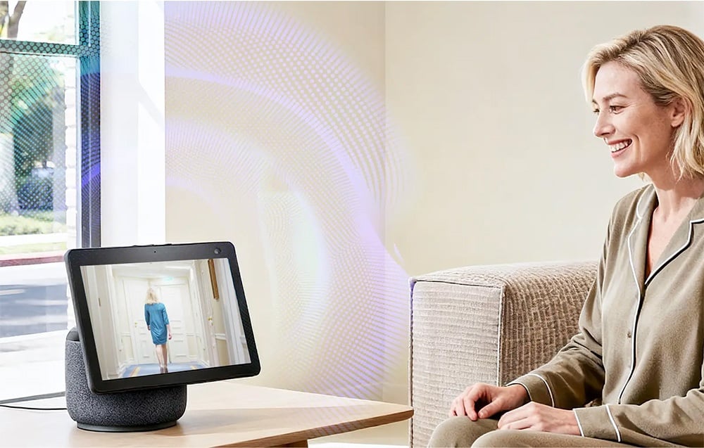 A woman sitting on a couch looks at a smart hub screen displaying a woman walking down a hallway