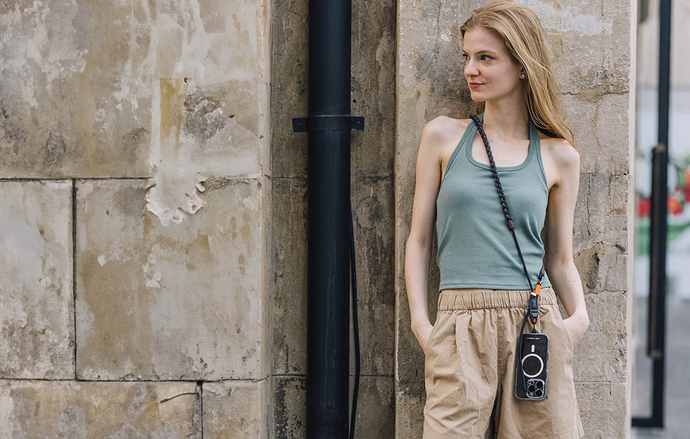 A woman leaning against a stone building wears a green T-shirt and a PGYTECH neck strap