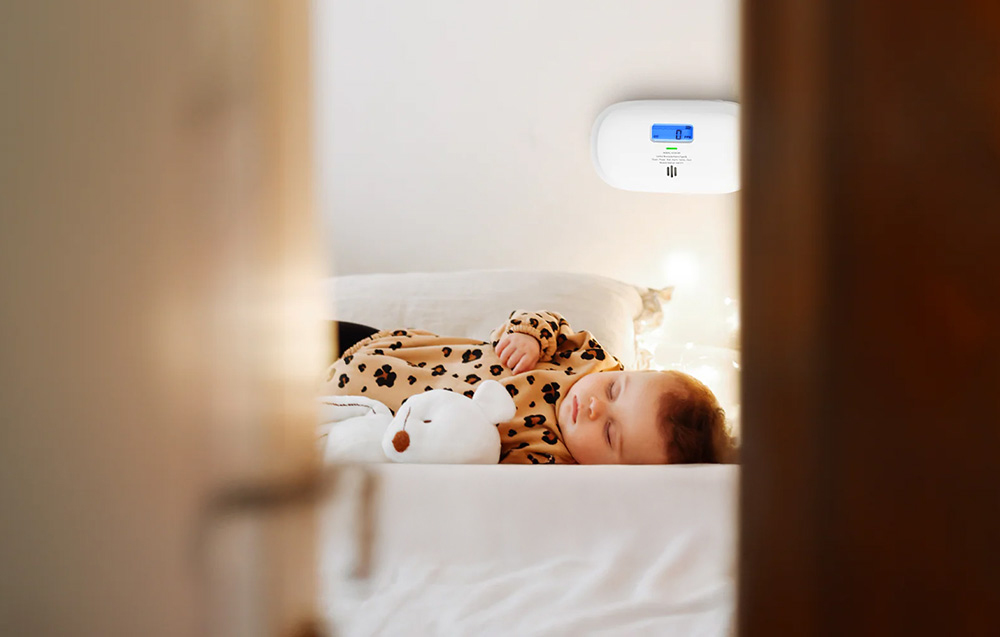 A sleeping child in bed with an X-Sense carbon monoxide detector mounted on the wall above it