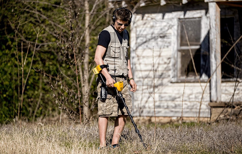 A man with a metal detector searches the ground near an abandoned house.
