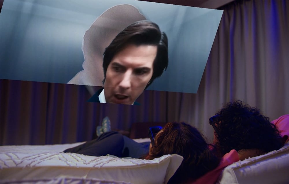 Two people lie on a bed and watch an image floating in the air, showing a close-up of the face of a man in a suit