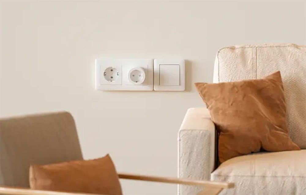 A white smart plug in a dual outlet on a light-colored wall behind a beige sofa with brown cushions