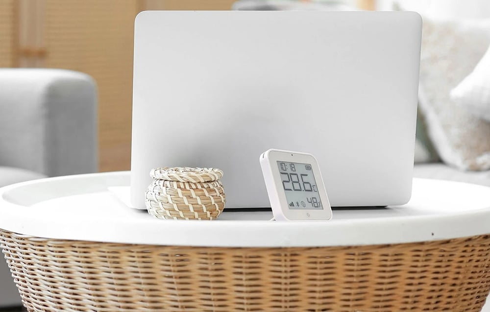 A white electronic temperature sensor standing next to a laptop on a wicker table in a bright living room