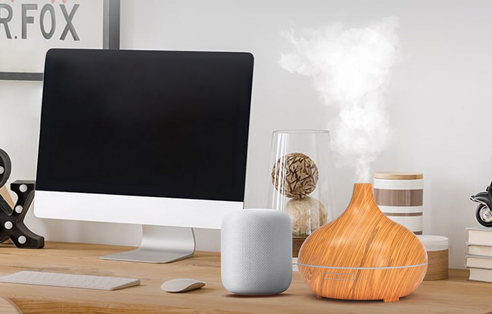 The oil diffuser in a wooden case stands on a desk next to an iMac and a white speaker