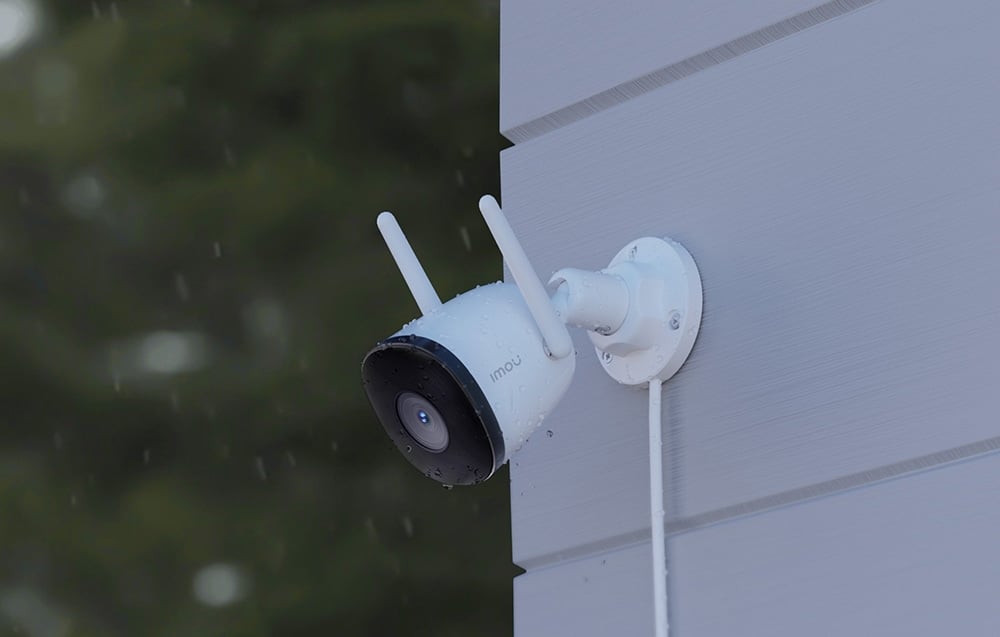 A white Imou camera with two antennas, mounted on a gray facade, covered with raindrops