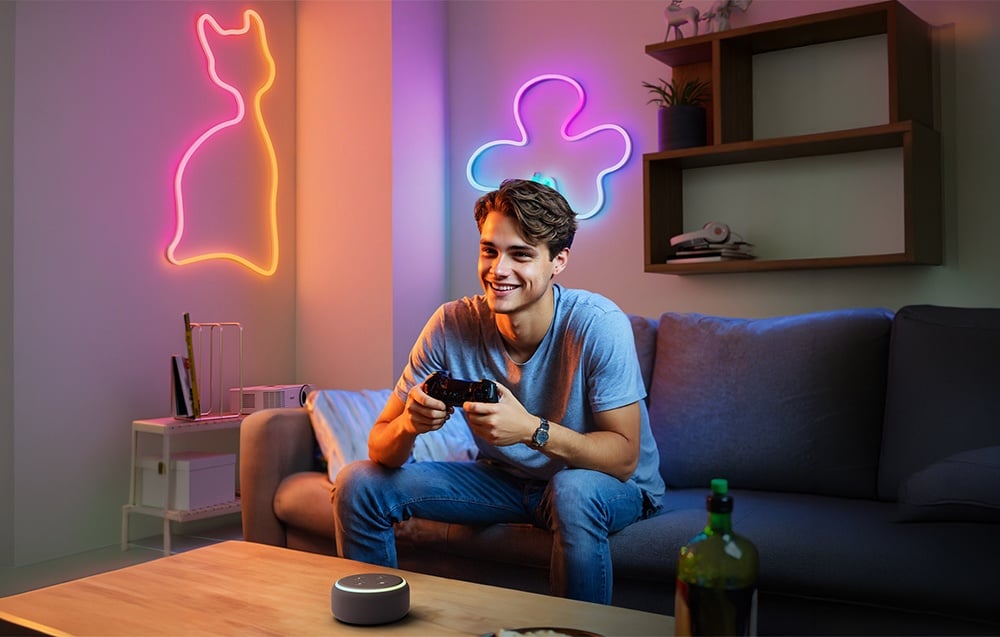 A gamer sits on the couch with a controller. Neon outlines of a cat and clouds glow on the walls.
