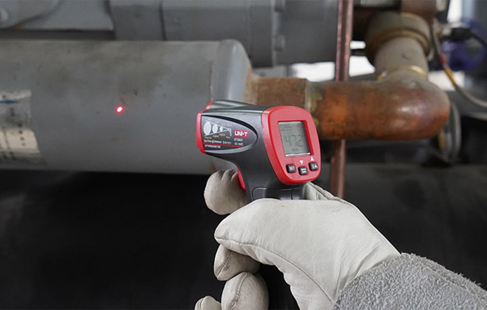 UNI-T pyrometer in a work glove during non-contact temperature measurement of an industrial pipe.