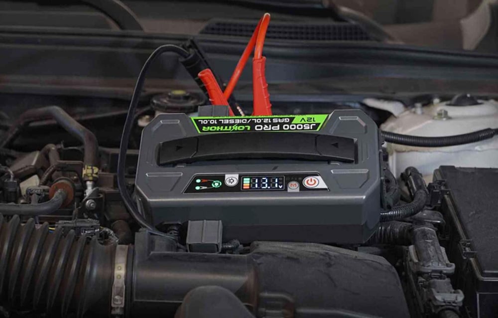 Portable jump starter connected with red clamps to the battery under the open hood of a car.