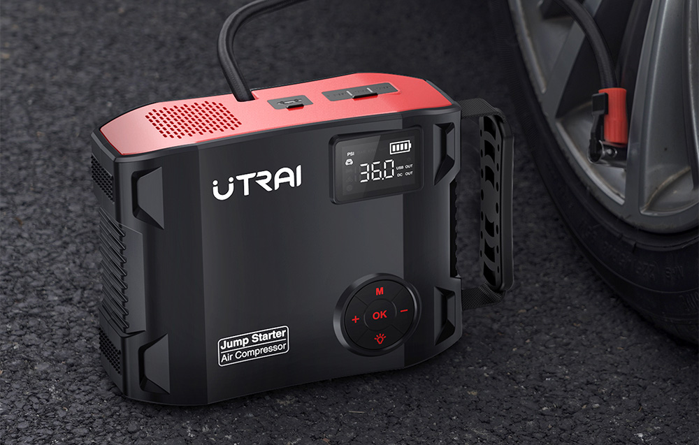 A black car jump starter sits on the asphalt and inflates a car tire using a built-in hose.