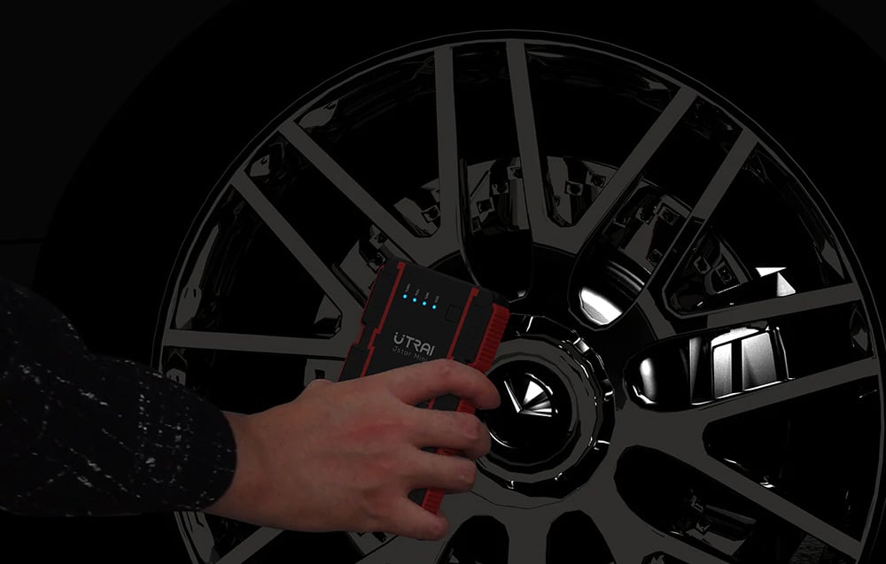 A man’s hand holding a black-and-red jump starter against the backdrop of a large car wheel.