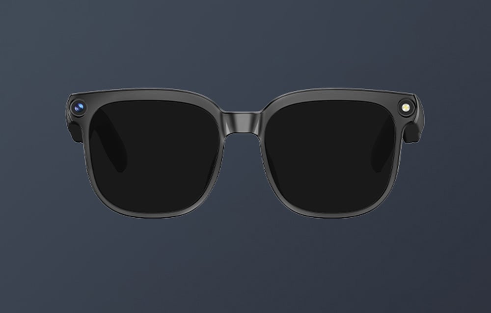 Front view of black smart glasses with a visible camera lens and sensors in the corners of the frames.