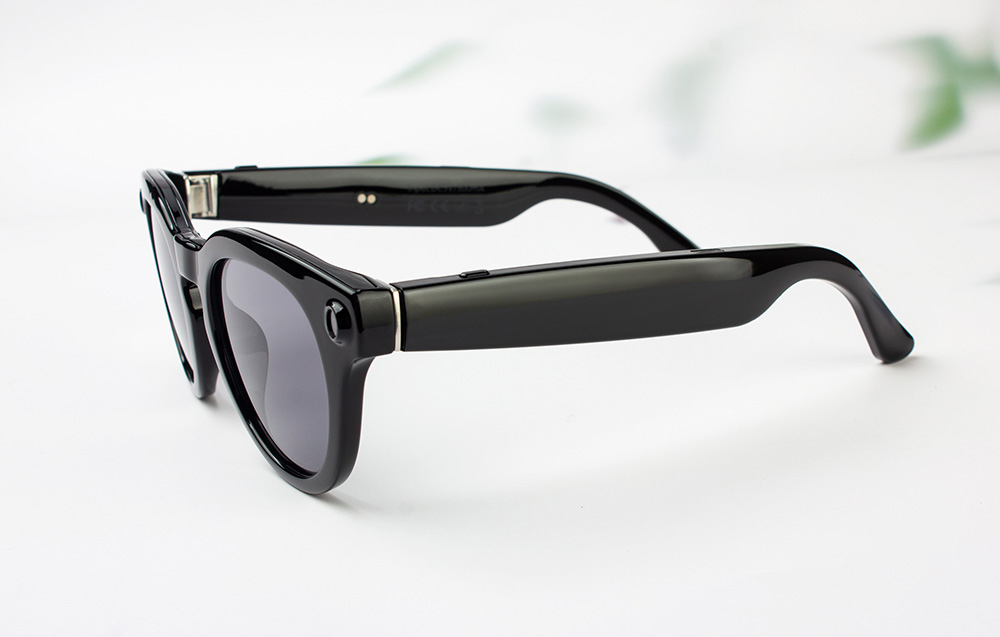 Product photo of black smart glasses with thick frames lying on a light-colored tabletop.