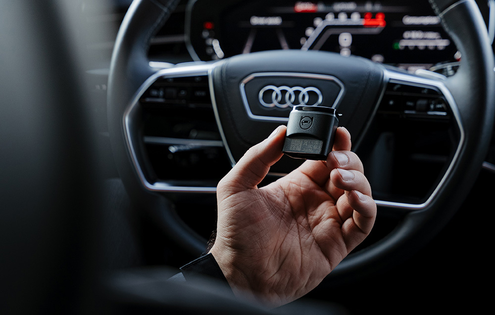 Carly OBD2 diagnostic adapter held in front of steering wheel with Audi logo in digital cockpit