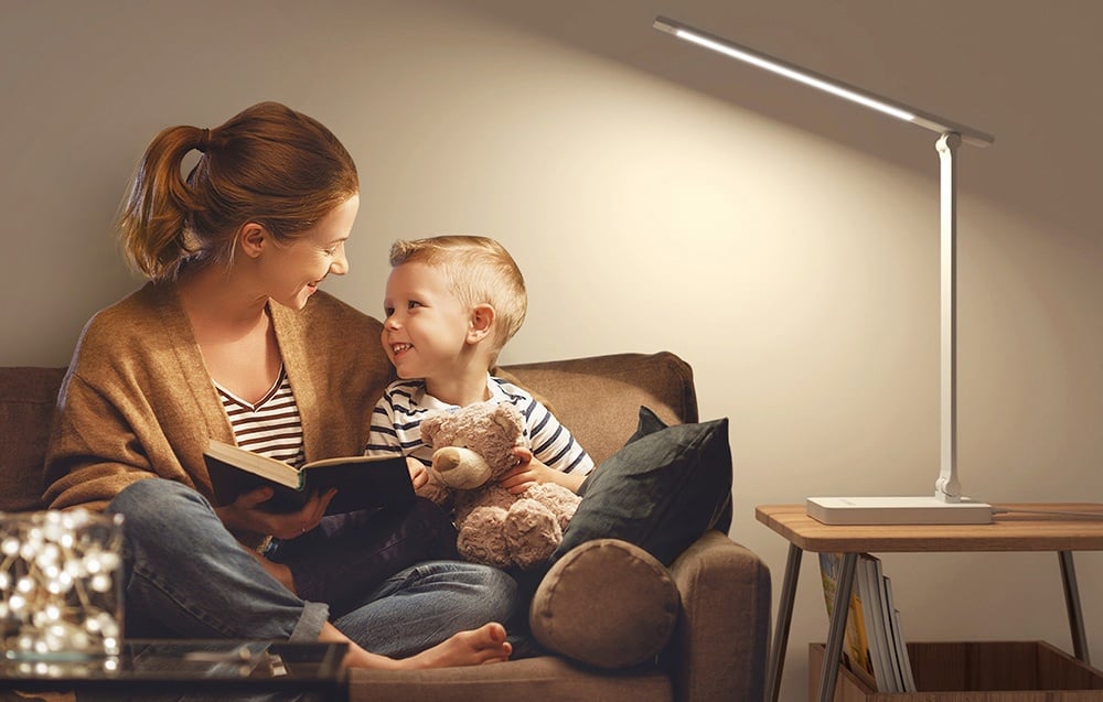 A woman reads a book to her child on the couch under the warm glow of the lamp standing nearby.