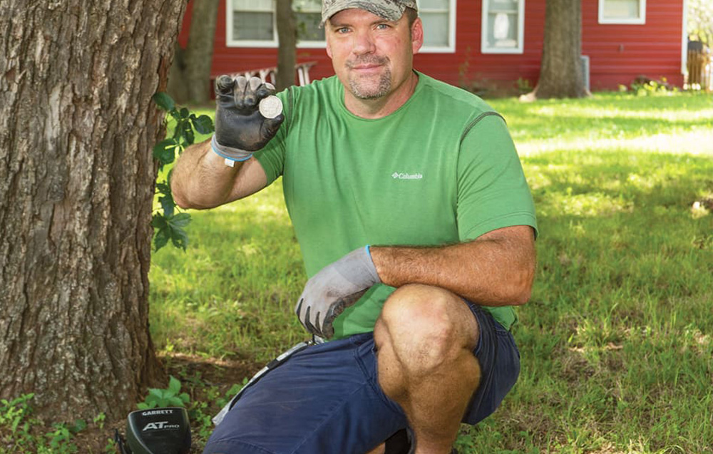 A man kneels on the grass and shows a coin found with a metal detector next to a tree.