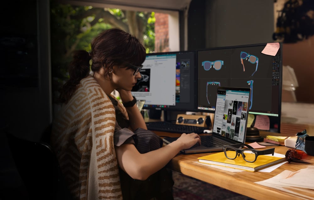 Woman working at a desk with three screens, with the glasses model lying next to a laptop.