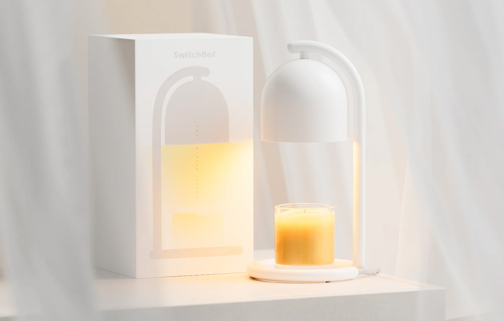 A white candle warmer heating a large yellow scented candle, placed next to a white packaging box on a light surface with a white background featuring a wavy fa