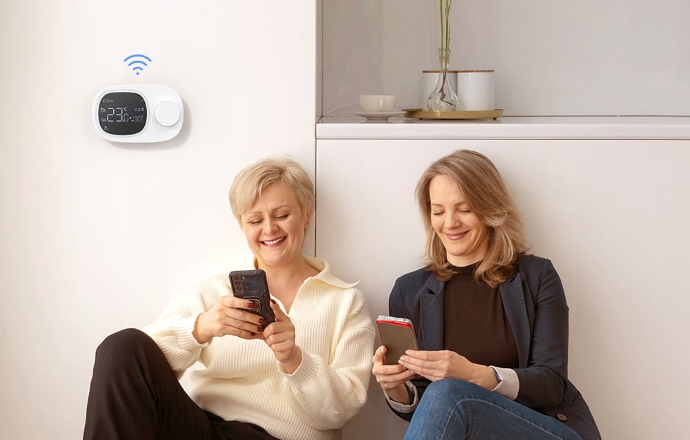 2 smiling women hold smartphones in their hands and sit on the floor. On the wall a smart thermostat.