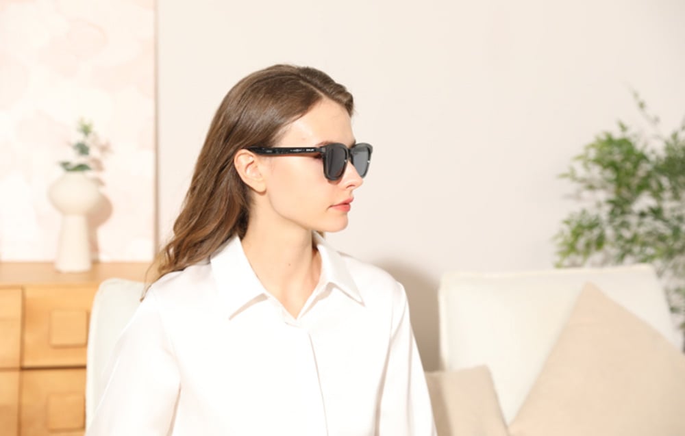 A woman sitting in a bright room, showing the side profile of smart glasses.
