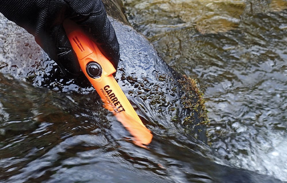A gloved hand dips a metal detector into a stream.