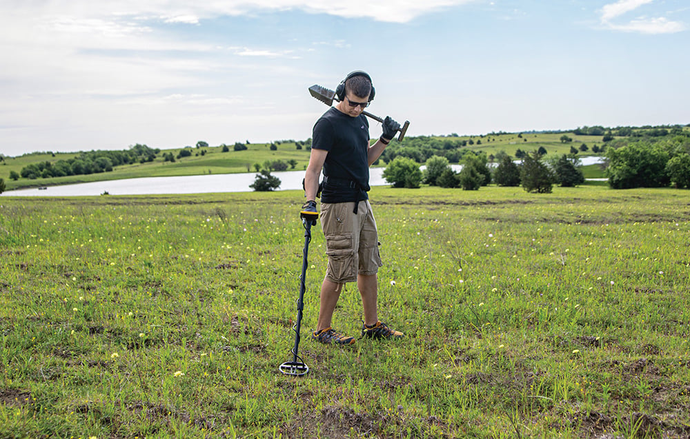 A man carries a metal detector on his shoulder against a green landscape.