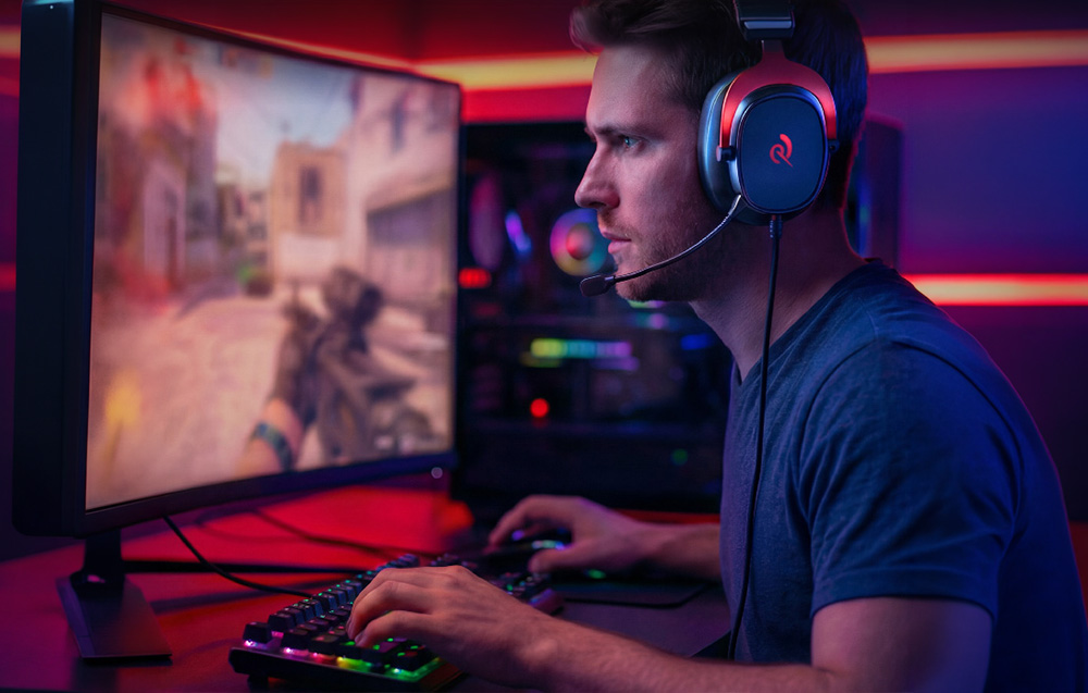 A man wearing gaming headphones playing an FPS shooter on a curved monitor in a room with red and blue lighting
