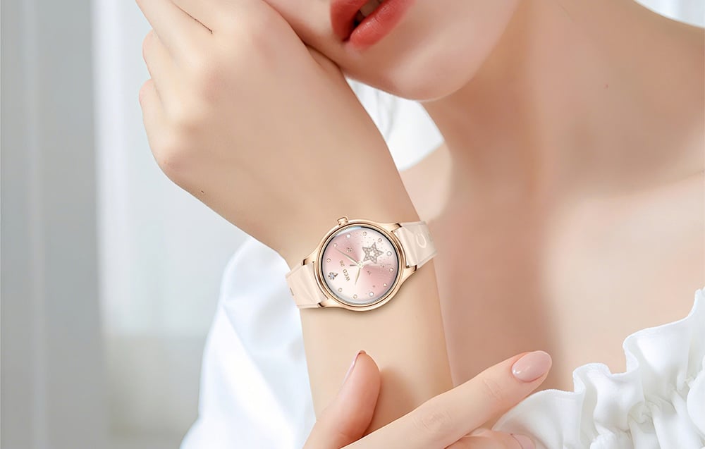 Close-up of a woman's wrist in a white blouse, presenting a gold smartwatch with a beige strap and a decorative pink dial with a star motif.