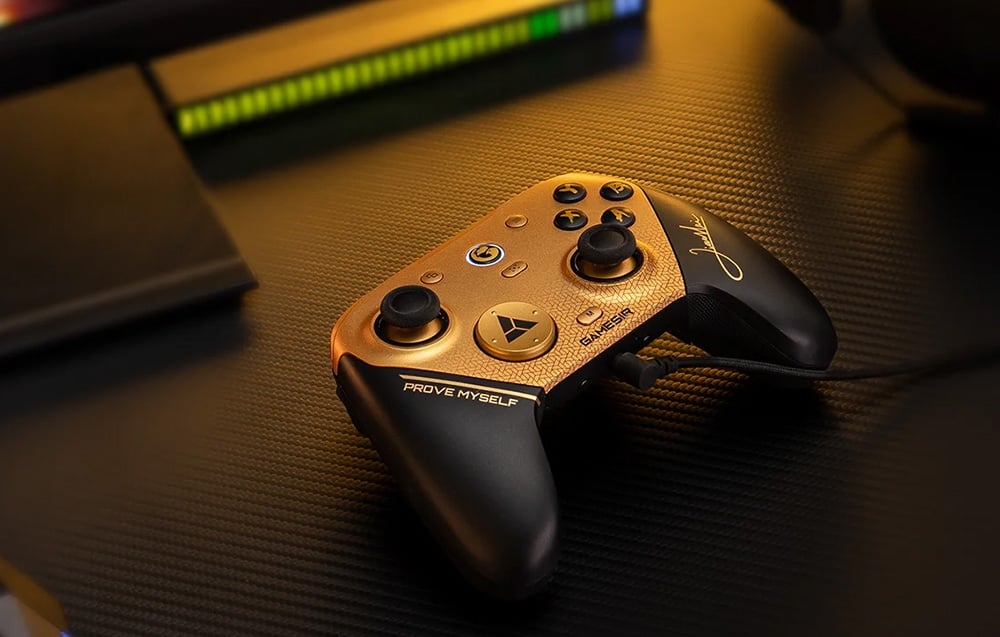 Gold-and-black game controller connected via a USB cable resting on a desk with backlighting in the background