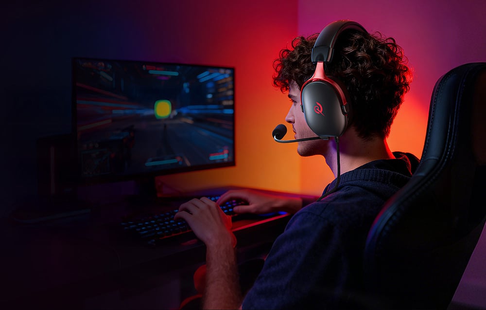 A man wearing headphones with a microphone playing a game on a computer in a dark room illuminated by colored light.