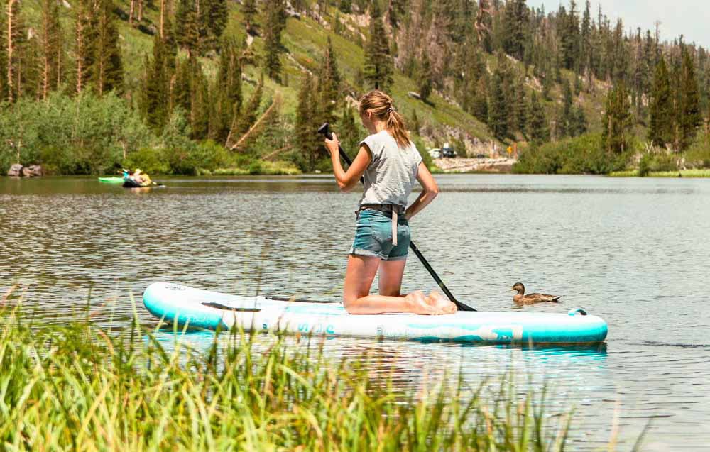 A woman with blonde hair on a surfboard. She is holding a paddle in her hand, with trees in the background