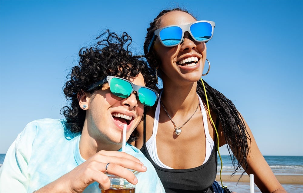Two smiling people wearing sunglasses with colored lenses are sitting on a beach under a blue sky