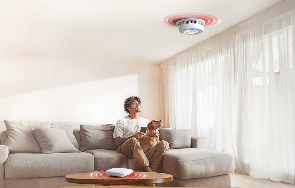 A man with a dog on the couch looks at a ceiling-mounted smoke detector communicating with a Wi-Fi gateway