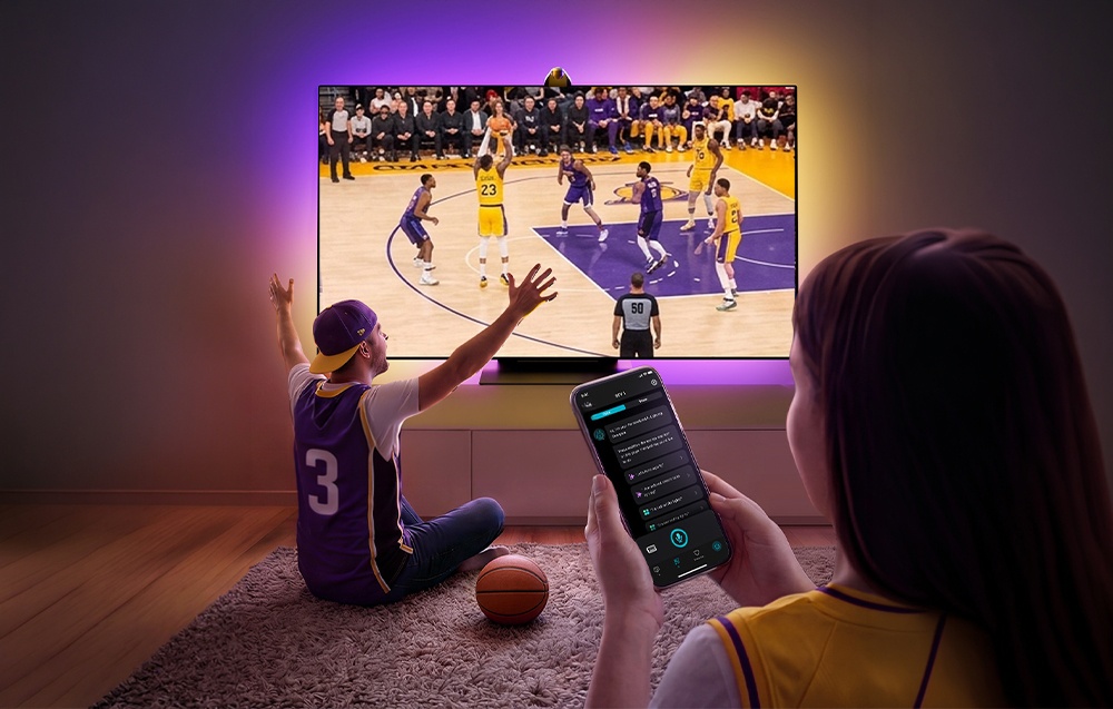 A woman holds a smartphone in her hand; a man sits on the carpet watching a basketball game; the wall behind the TV is lit in purple and yellow