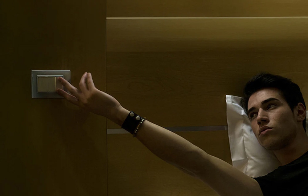 A person lying in bed reaches out to a light switch on a wooden wall