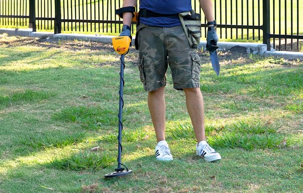 Person with metal detector standing on grass, holding shovel.