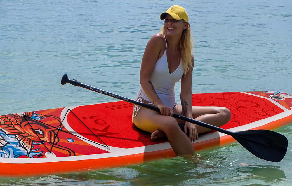 A smiling woman wearing a yellow baseball cap is sitting on a red swimming board in the water. She is holding a paddle in her hand
