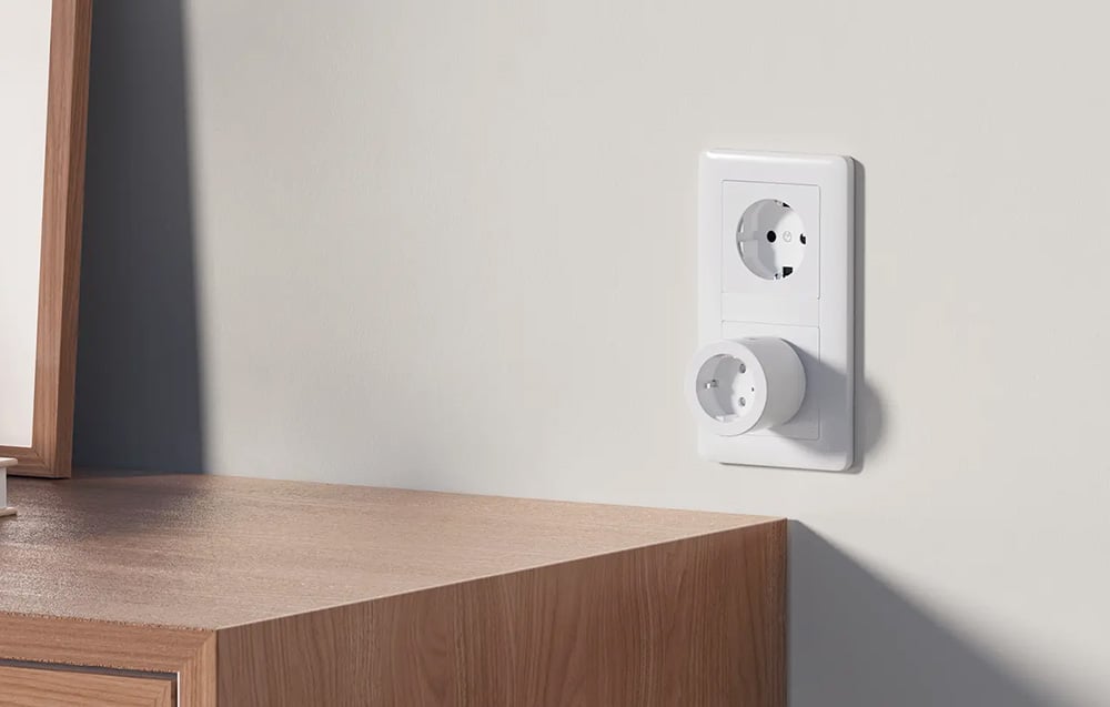 A white smart plug plugged into a vertical wall outlet above a wooden cabinet.