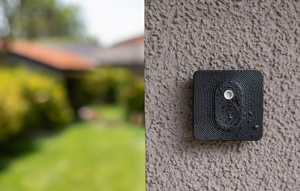 A black Shelly button covered in water droplets mounted on a rough exterior wall
