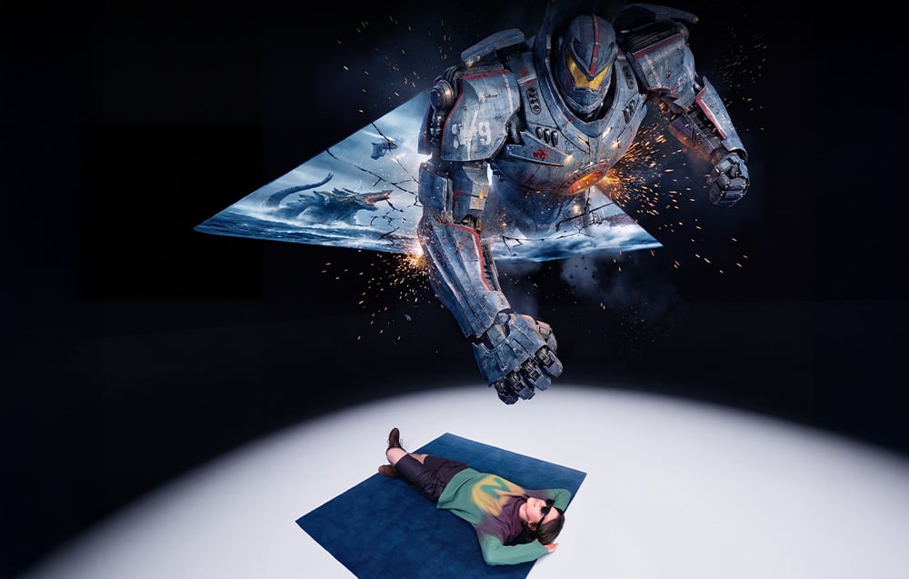 A smiling man lies on a blanket wearing AR glasses that display a character from a game stepping out of the screen