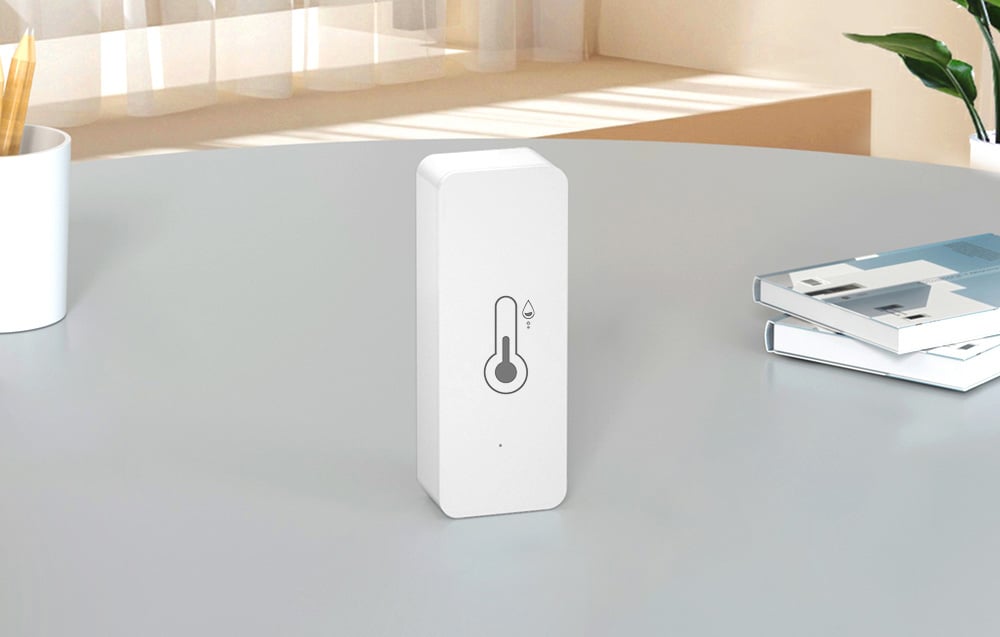 White temperature and humidity sensor with a thermometer icon on the front, set on a gray table in a bright room
