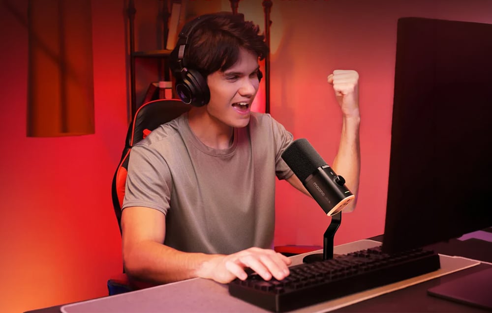 A smiling man wearing headphones in front of a computer, making a victory gesture next to the Maono microphone