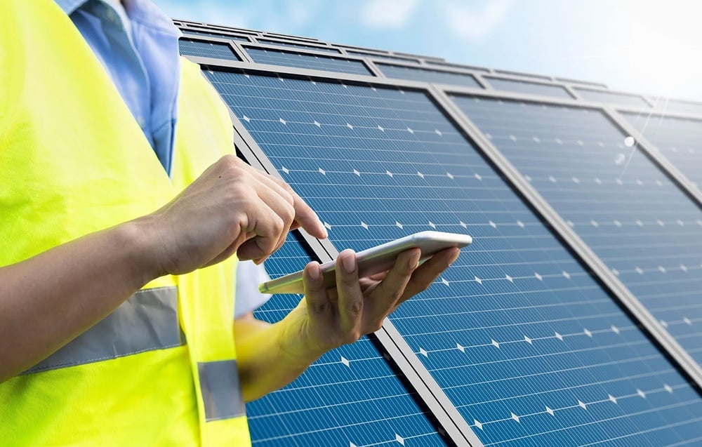 A person in a yellow reflective vest holding a smartphone against a backdrop of solar panels