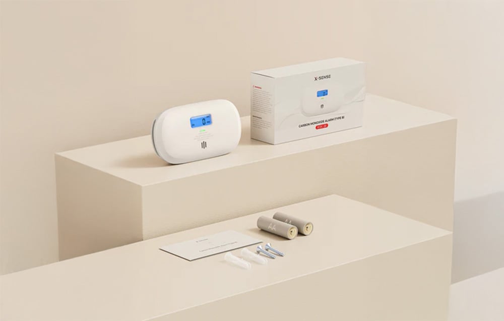 X-Sense carbon monoxide detector with LCD display on platform, next to packaging, batteries, instructions and mounting accessories