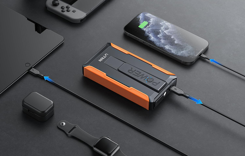 A multifunctional car starter charged via a cable, which is powering a cell phone lying next to it.