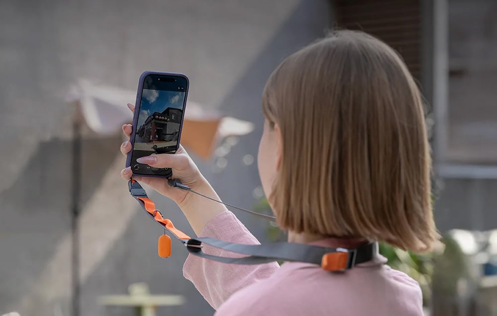 A woman with short hair photographs a building with her phone attached to a neck strap