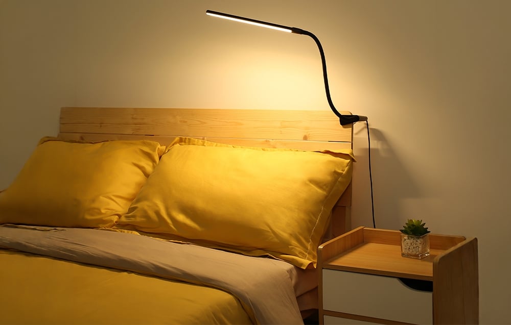 A desk lamp with a flexible arm clamped to a wooden headboard, casting a warm glow over yellow pillows and a comforter.