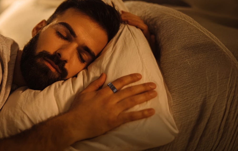 A bearded man sleeps on a pillow with a black smart ring on his finger in warm, homey lighting