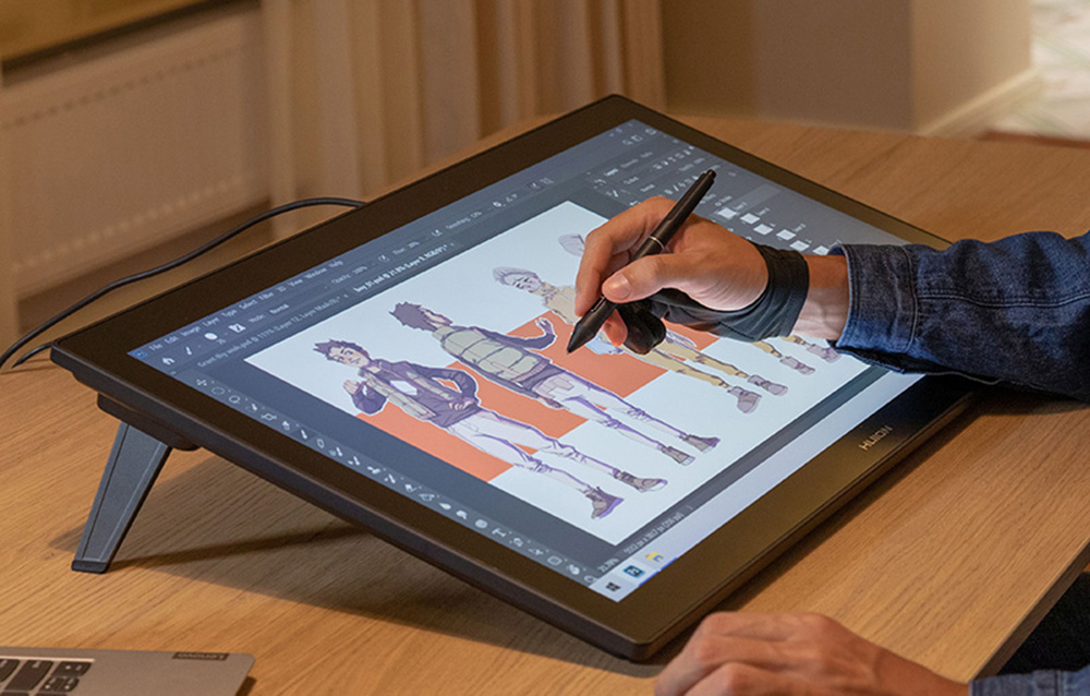 Huion tablet on desk with stand, person drawing characters in graphics program, laptop in background