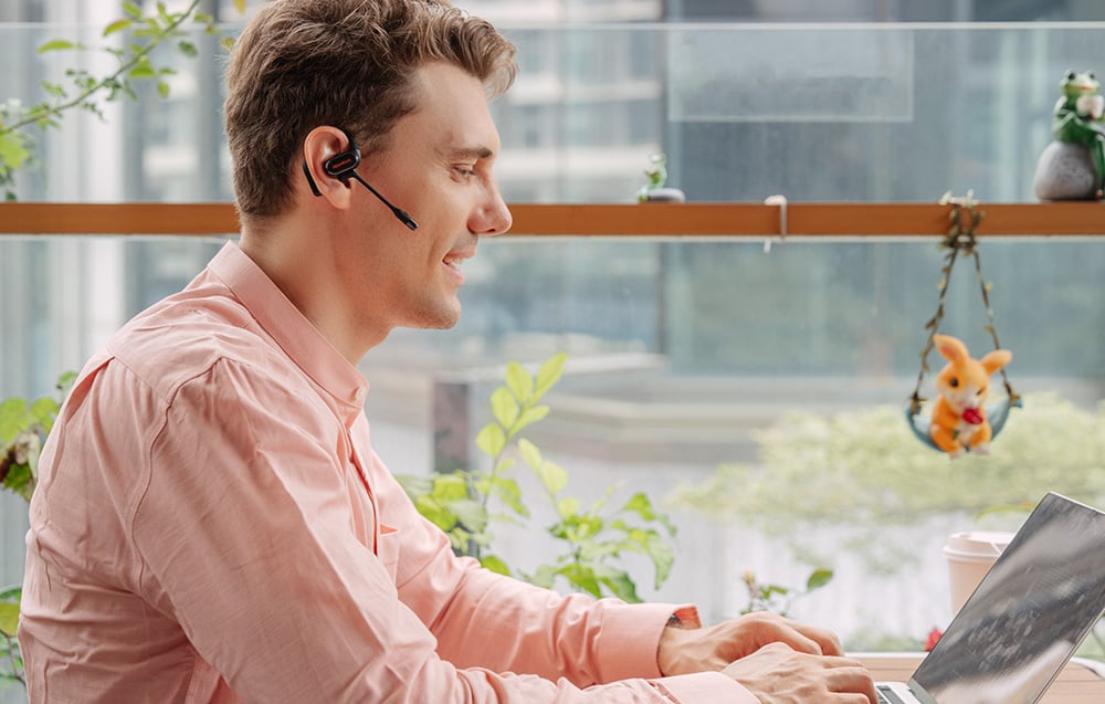 Man in pink shirt working at laptop, wearing black headphones with microphone
